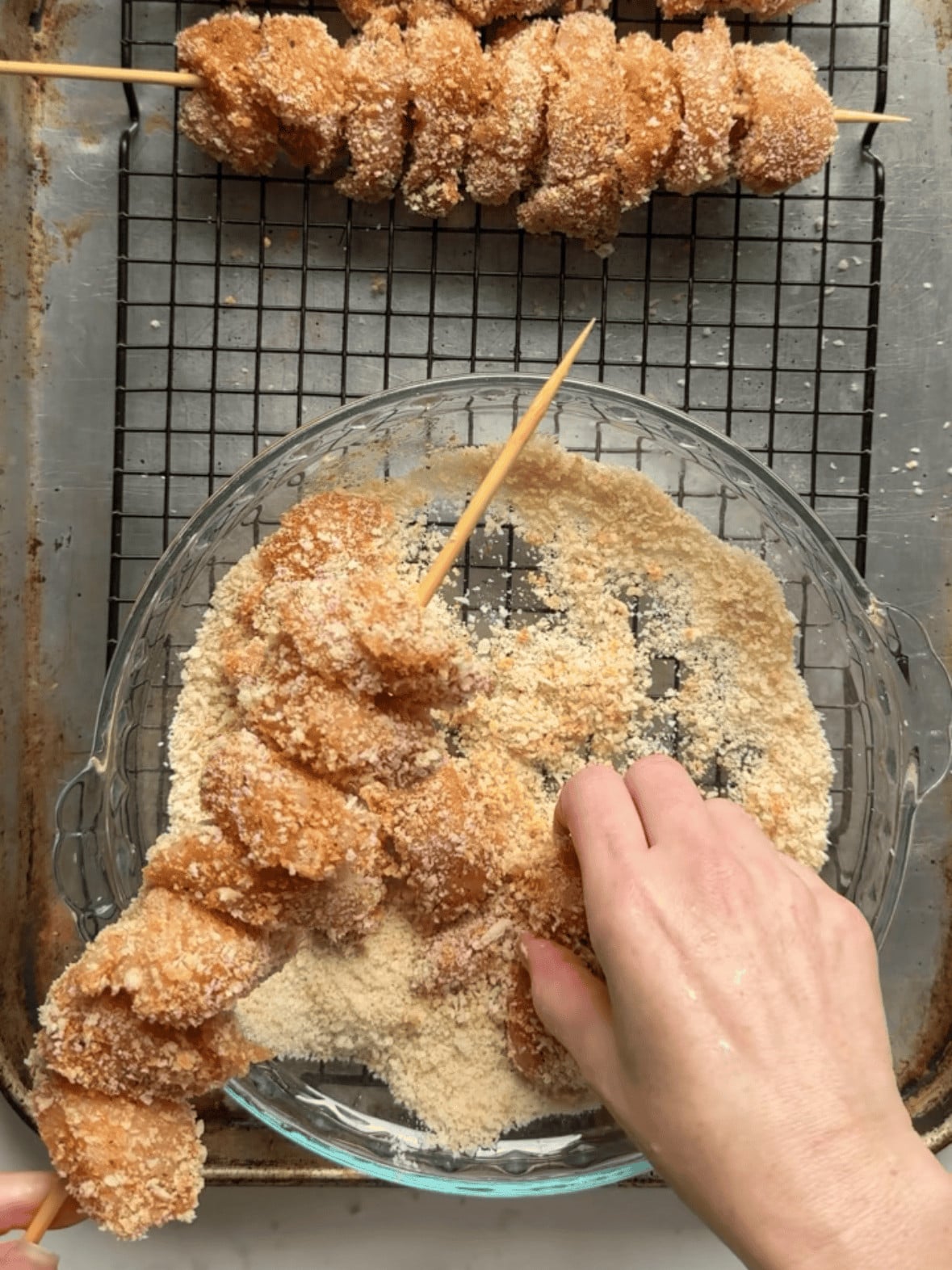 Overhead view of hands pressing Panko breaded chicken cubes onto a wooden skewer over a wire rack on a baking sheet, with completed skewers lined up in the background ready for the oven