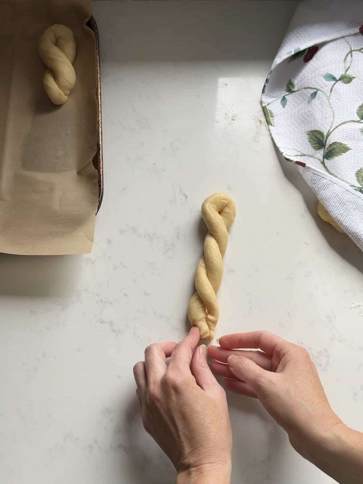 Hands twisting cheoreg dough into shape on a marble counter with a baking sheet in the background