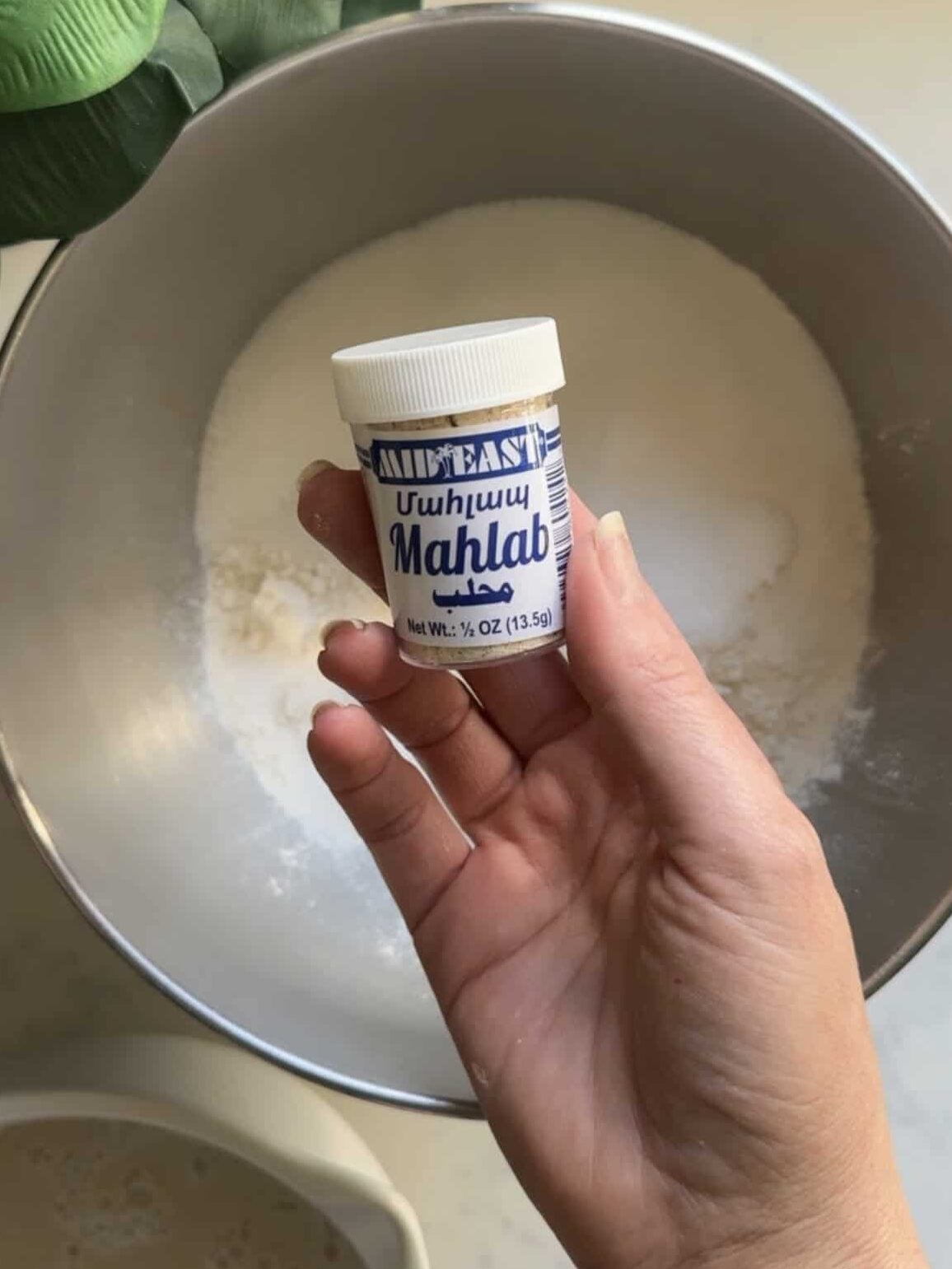 Hand holding a small jar of Mid East Mahlab mahleb spice over a stand mixer bowl with flour for cheoreg Armenian sweet bread