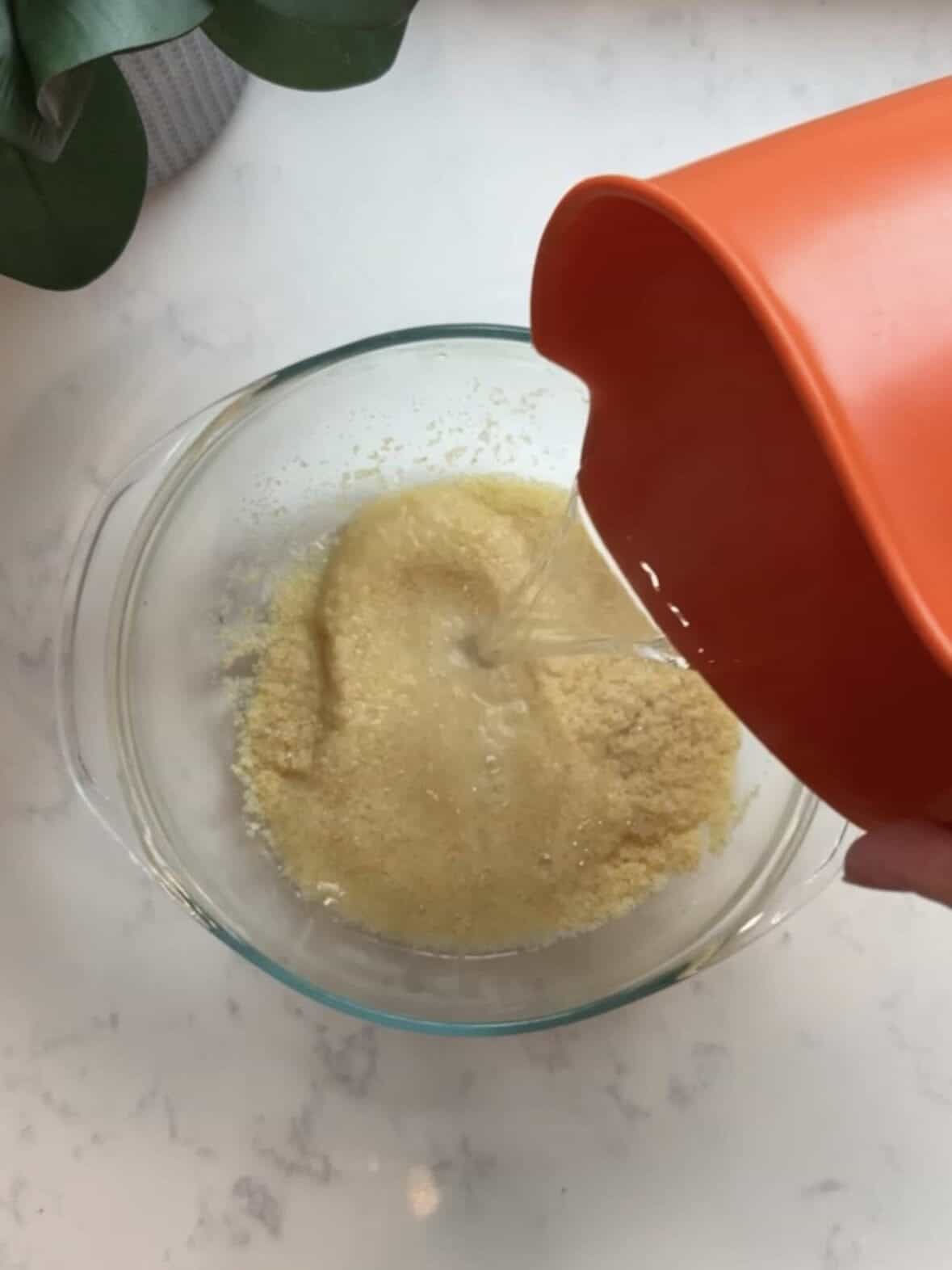 Boiling water being poured from an orange measuring cup over fine bulgur in a glass bowl on a marble countertop