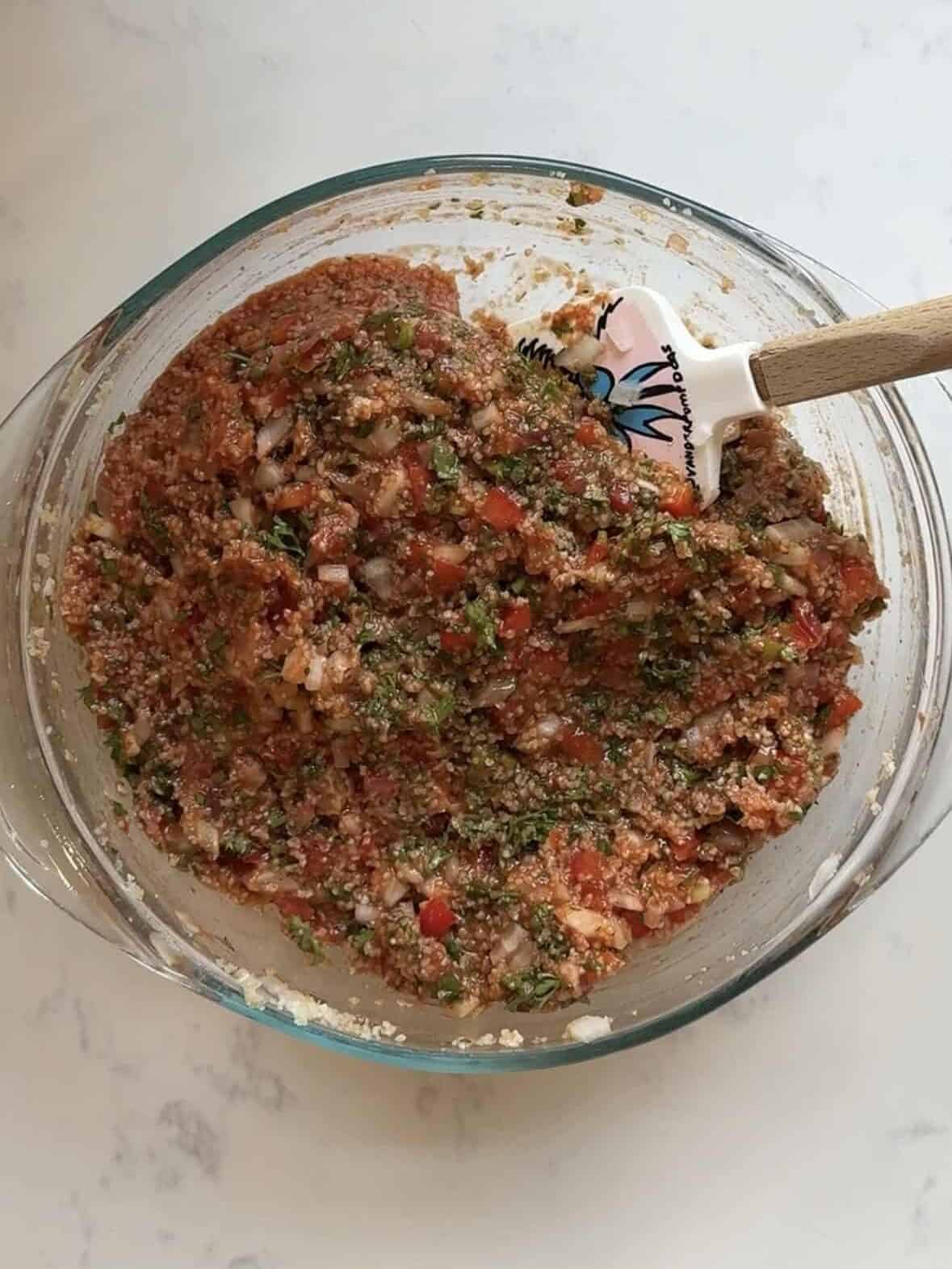Fully mixed Armenian eech red tabouli in a glass bowl on a marble countertop with a wooden handled spatula
