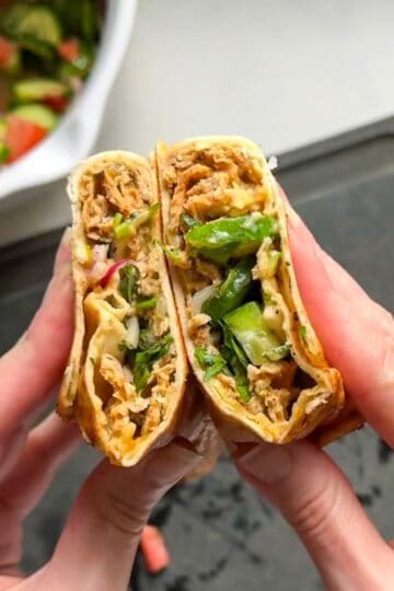 Hands holding a sliced chicken doner kabob wrap showing layers of spiced ground chicken hummus and fresh cucumber salad inside golden toasted flatbread