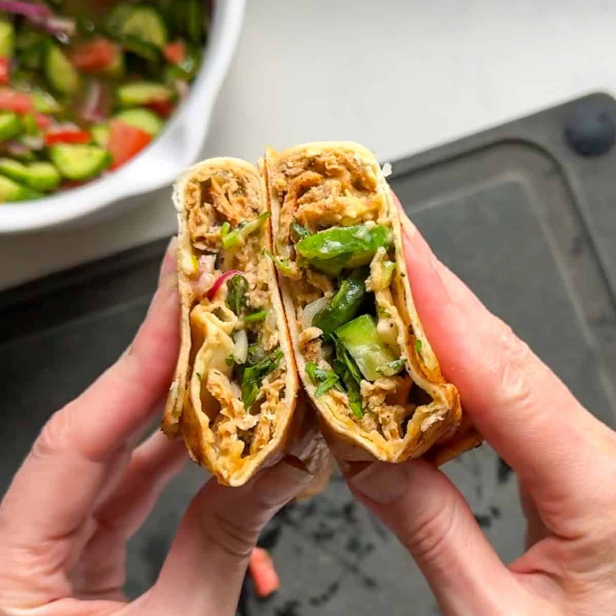Hands holding a sliced chicken doner kabob wrap showing layers of spiced ground chicken hummus and fresh cucumber salad inside golden toasted flatbread