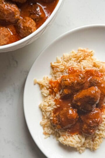 Overhead view of sujakaki soutzoukakia Smyrna meatballs in tomato sauce served over rice pilaf on a white plate with serving bowl on white marble surface