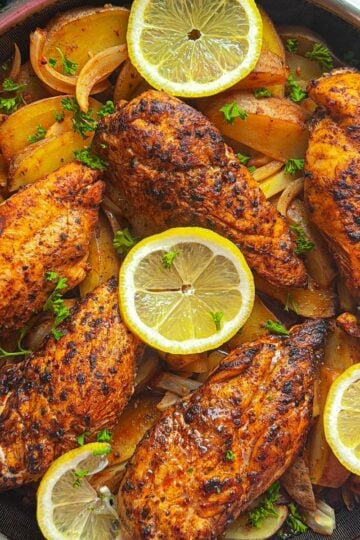 Overhead view of Aleppo pepper chicken breasts and potatoes in a dark skillet garnished with lemon slices and fresh parsley