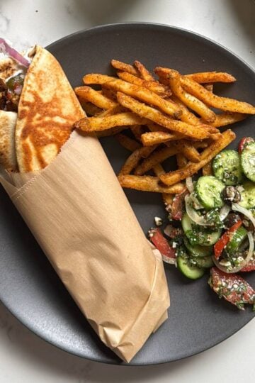 Close up of a chicken shawarma wrap in parchment paper being held over a plate with seasoned fries and cucumber salad