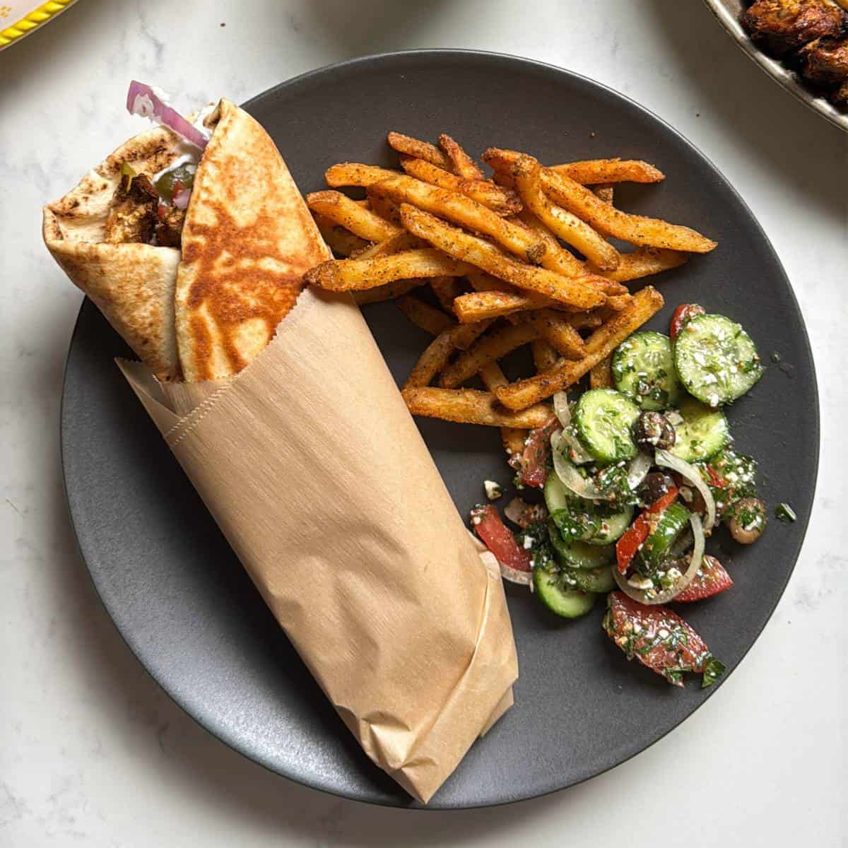 Close up of a chicken shawarma wrap in parchment paper being held over a plate with seasoned fries and cucumber salad