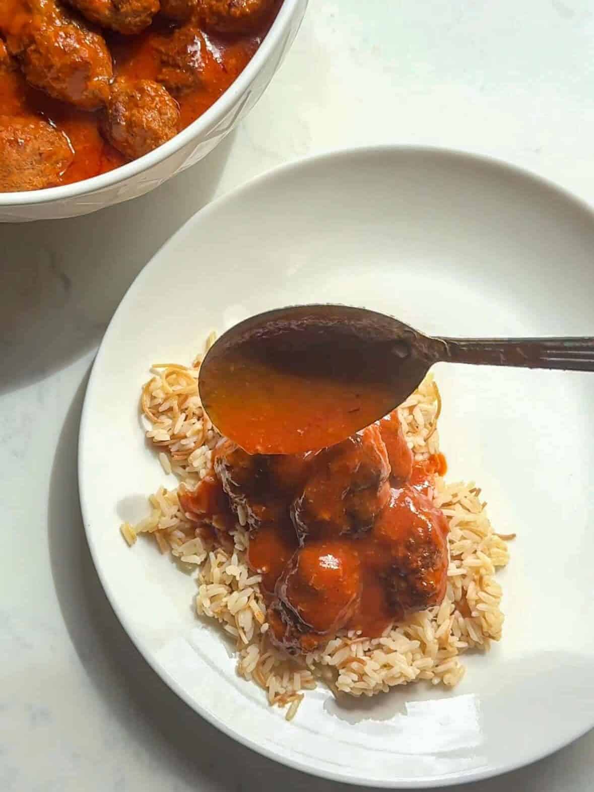 Armenian rice pilaf topped with sujakaki meatballs in red tomato sauce on a white plate, a spoon drizzling sauce over the top