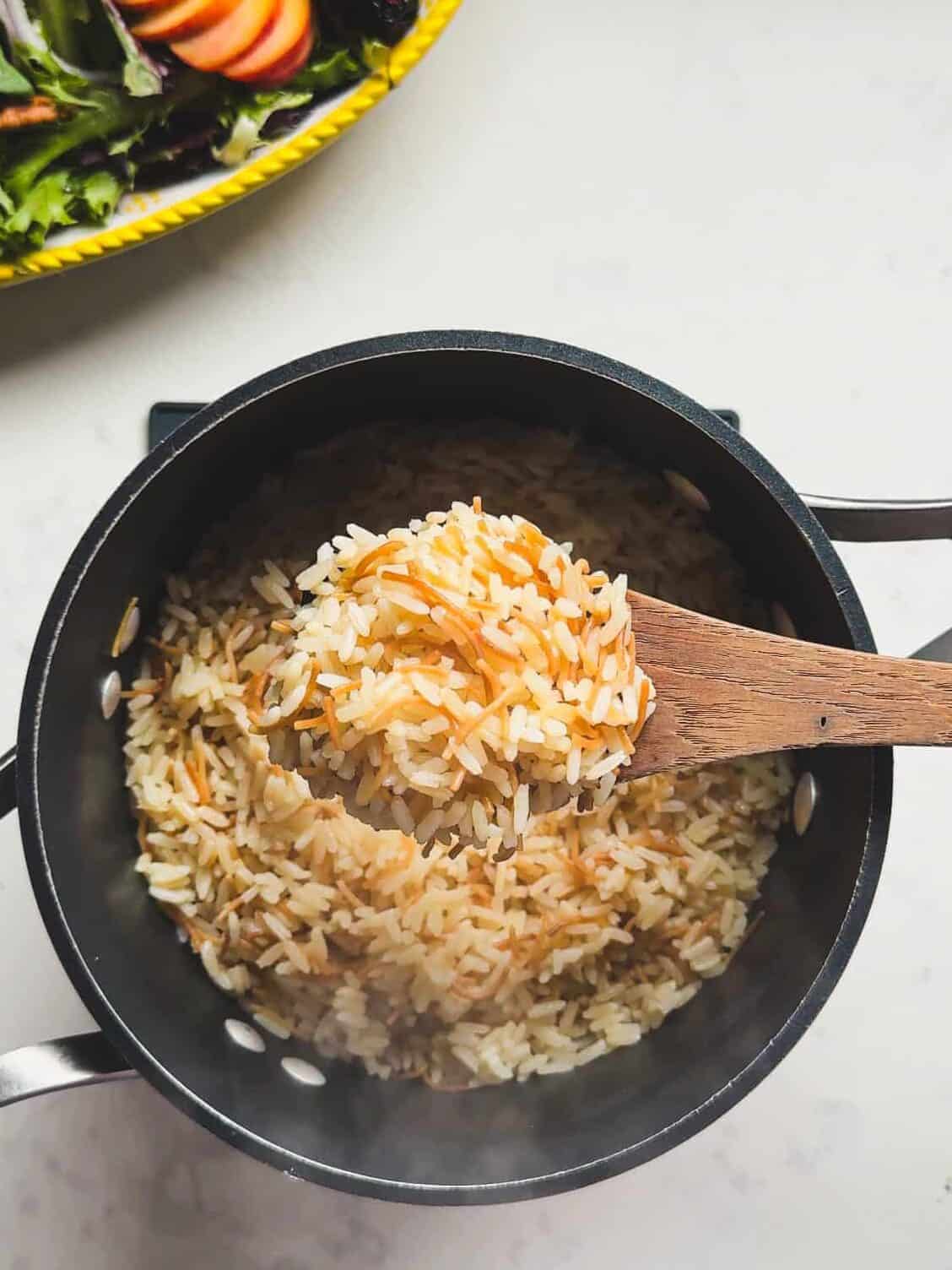 Overhead view of a wooden spoon scooping fluffy Armenian rice pilaf with toasted noodles from a pot