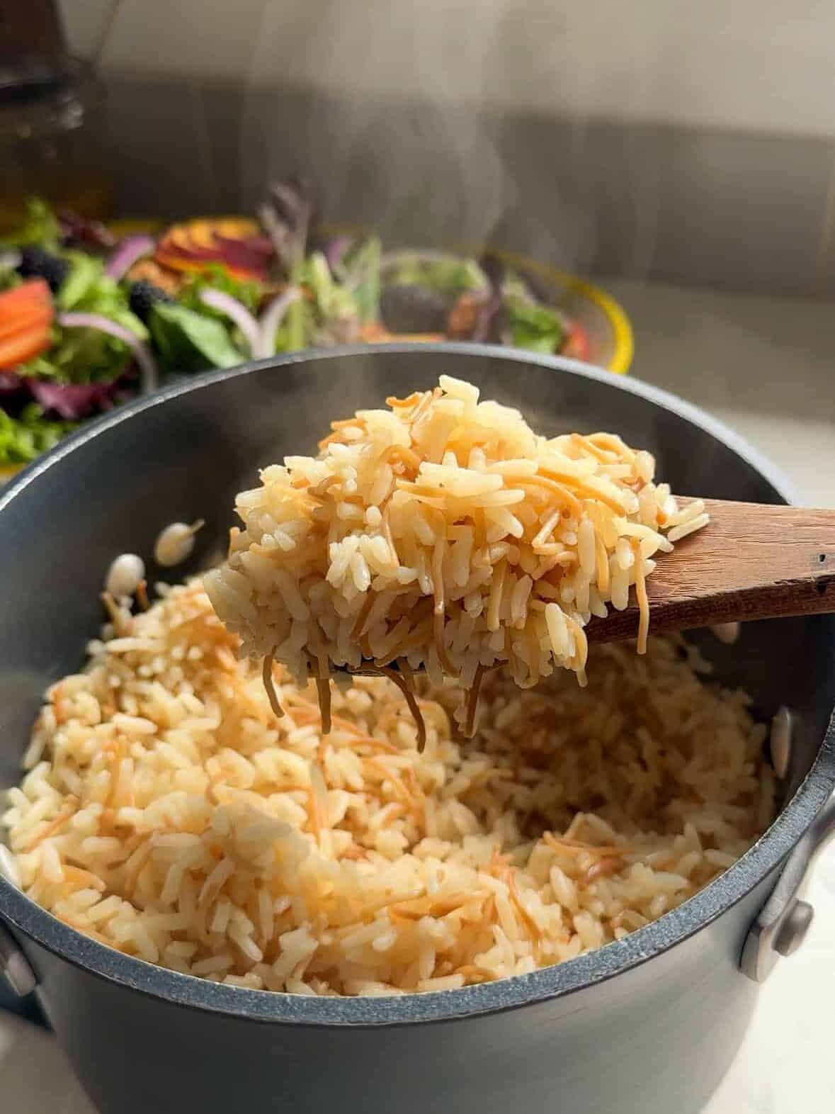Steaming spoonful of fluffy Armenian rice pilaf with toasted noodles held above the pot, fresh salad behind