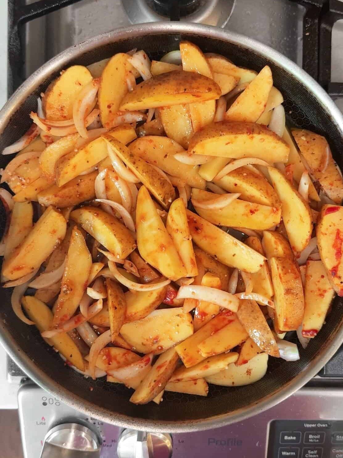 Yellow potato wedges and sliced onions coated in Aleppo pepper seasoning cooking in a large skillet on a gas stovetop