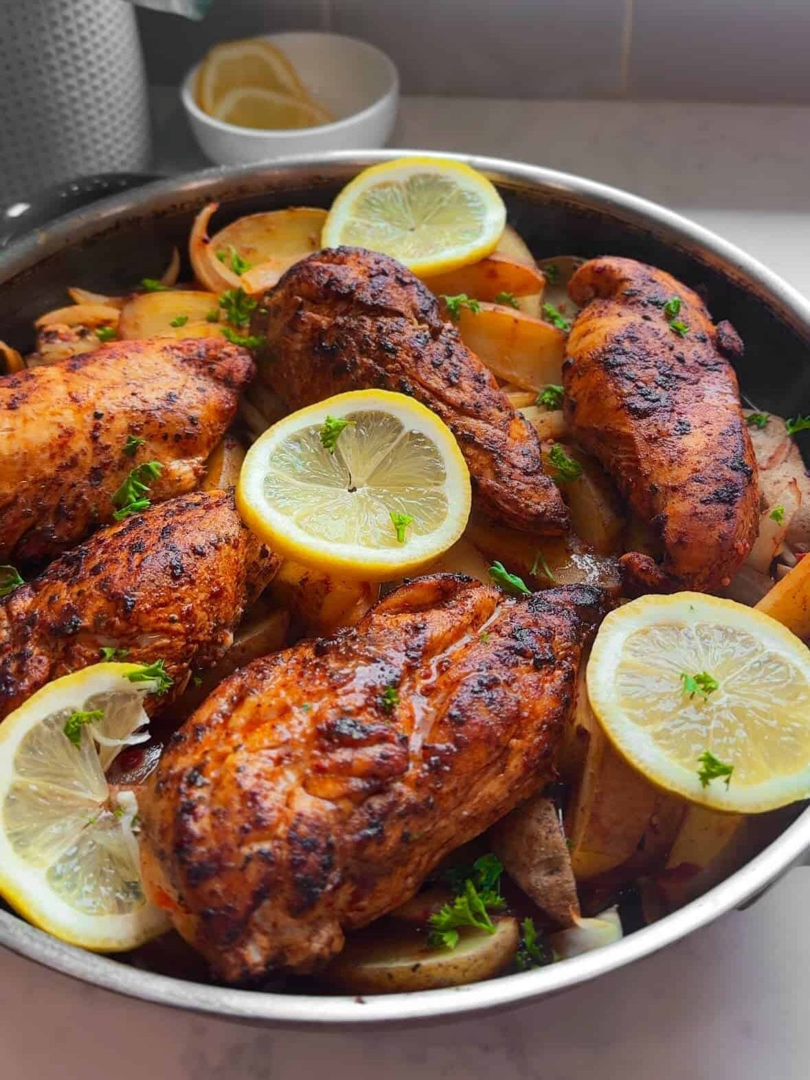 Aleppo pepper chicken and potatoes in a silver skillet with lemon slices and parsley near a kitchen window
