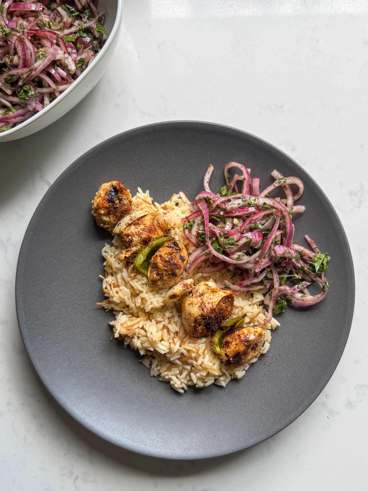 Plate of grilled chicken kabobs, rice pilaf, and sumac salad with the salad bowl in the background