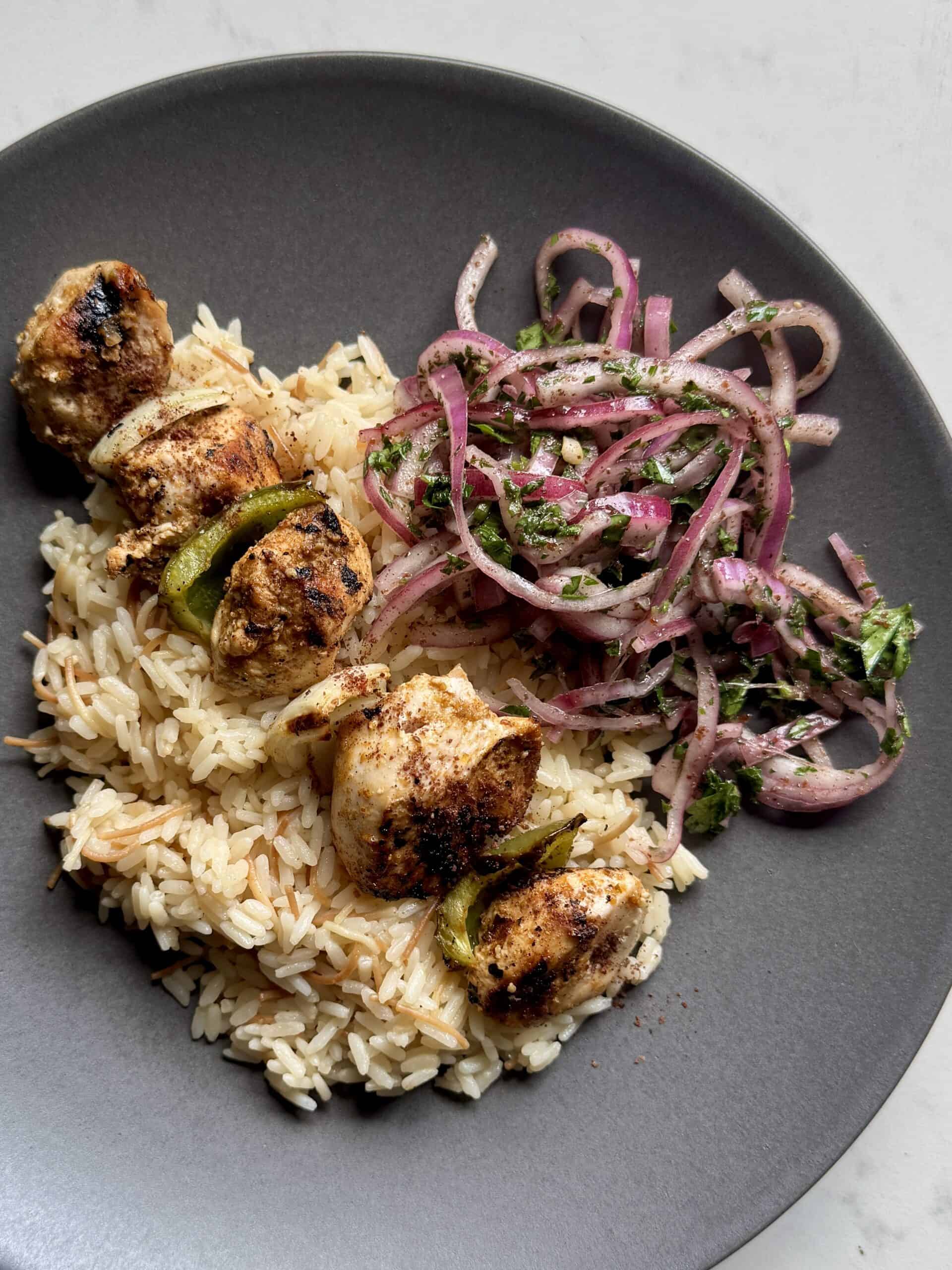 Close-up of a dinner plate with chicken kabobs, rice pilaf, and sumac onion salad