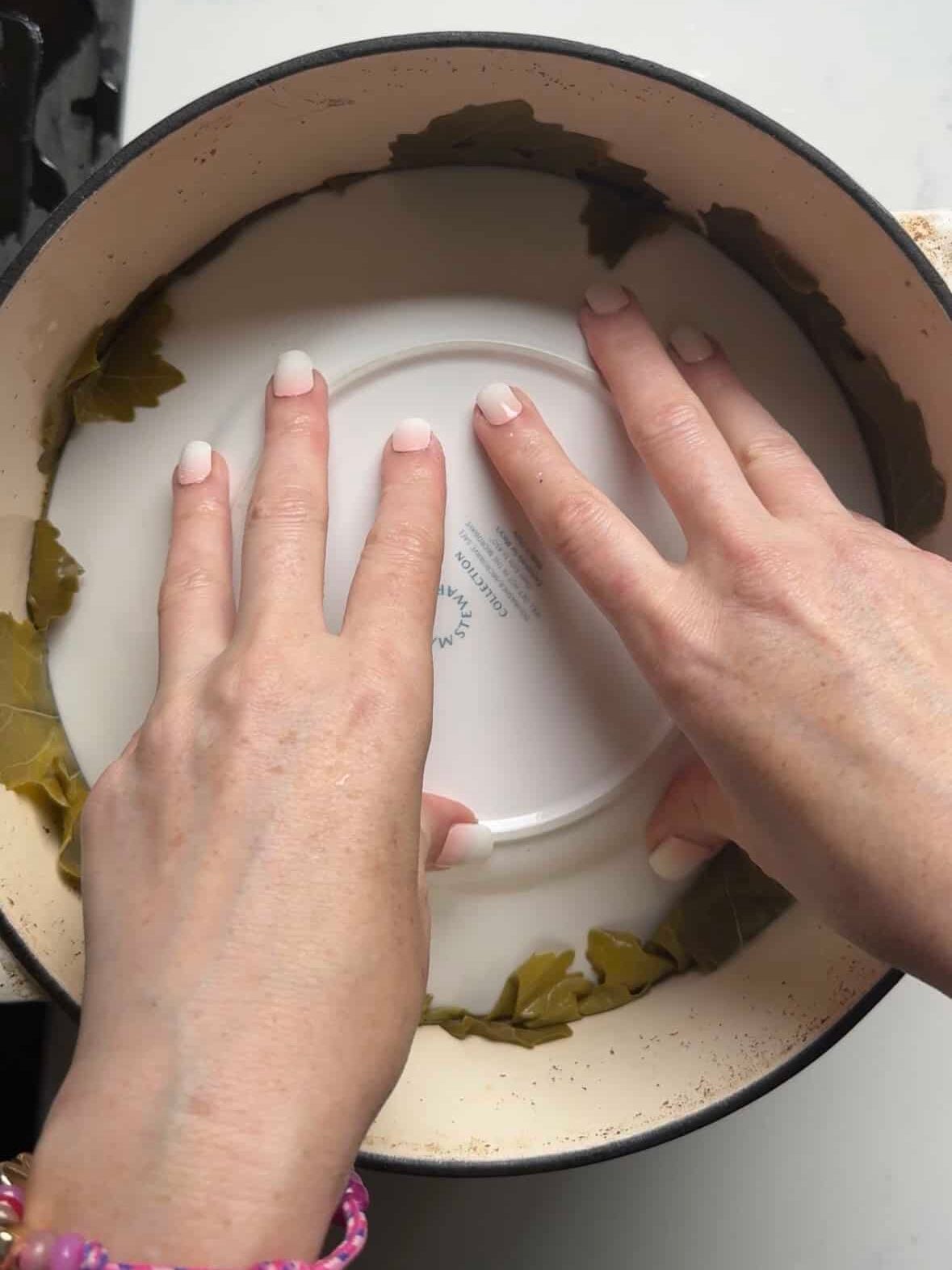 Hands pressing an inverted plate over grape leaves in a Dutch oven