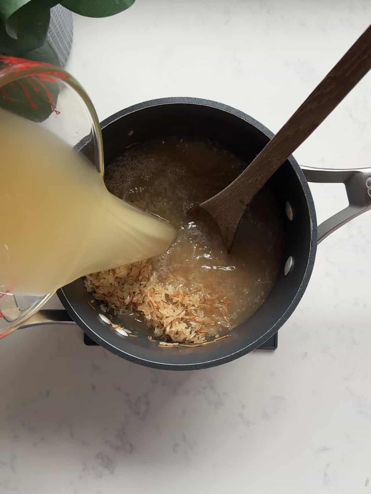 Chicken broth being poured from a measuring cup into a pot of toasted rice and noodles for rice pilaf