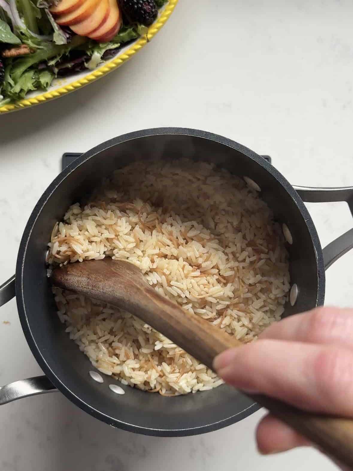 Wooden spoon fluffing finished Armenian rice pilaf in a pot, fresh salad in the background