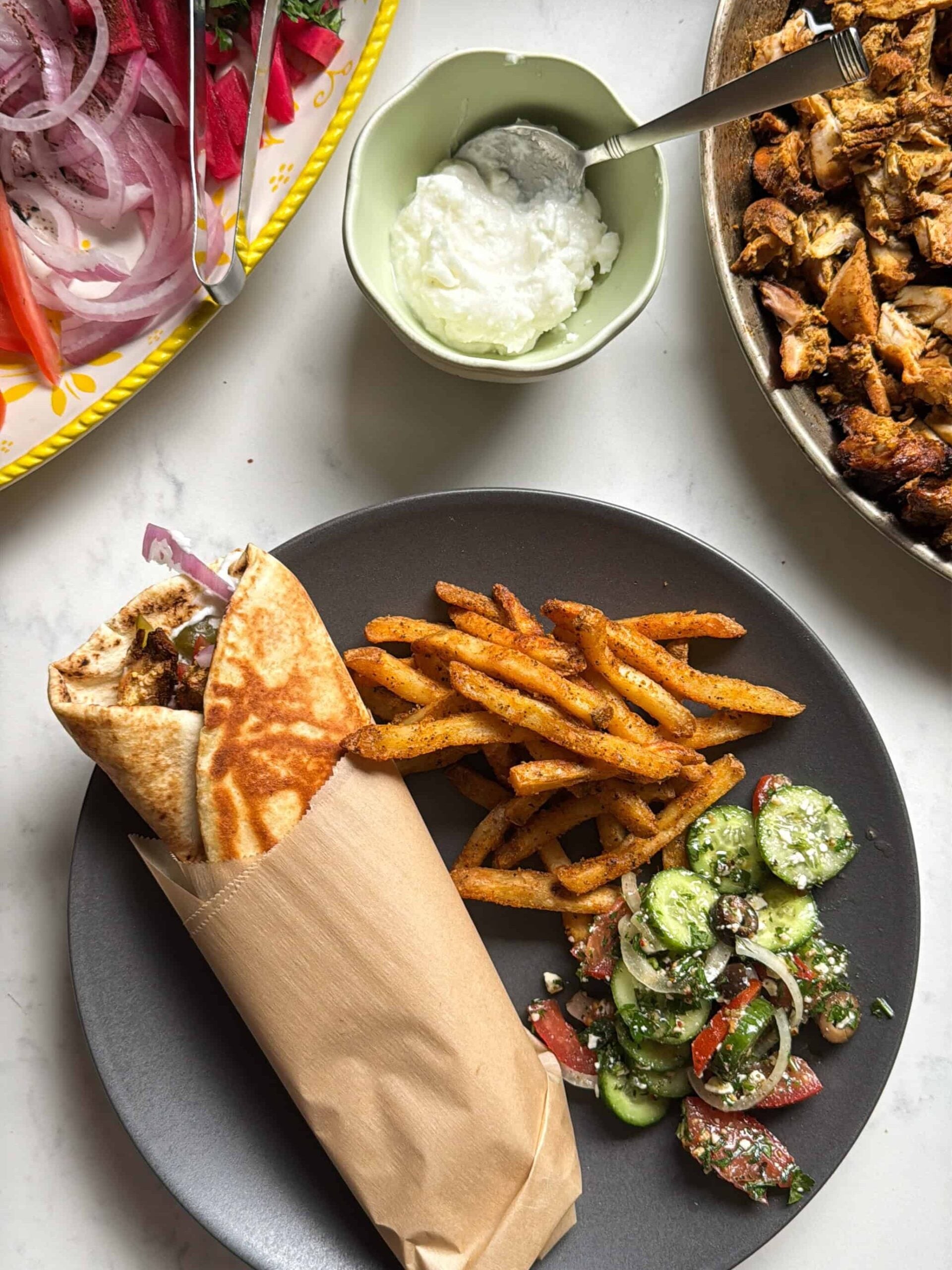 Finished chicken shawarma wrap wrapped in parchment paper served with seasoned fries and cucumber tomato salad