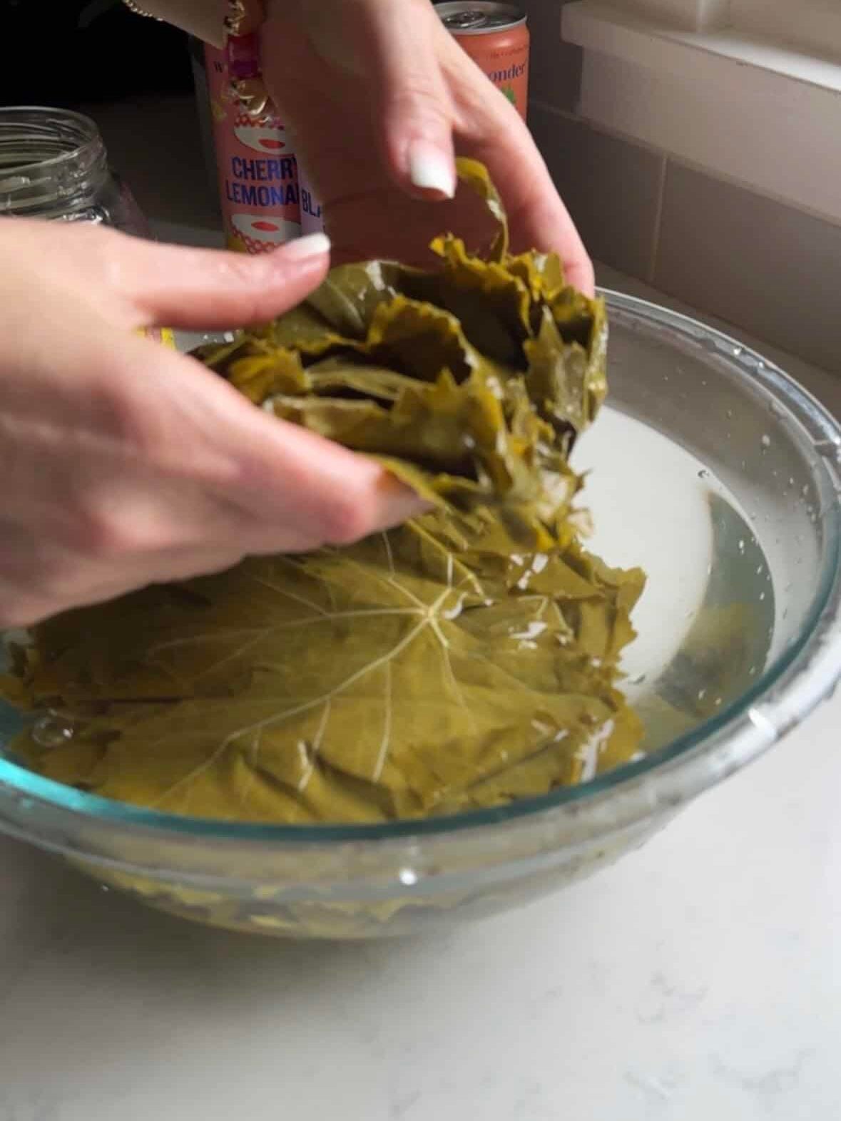 Hands rinsing brined grape leaves in a glass bowl of water