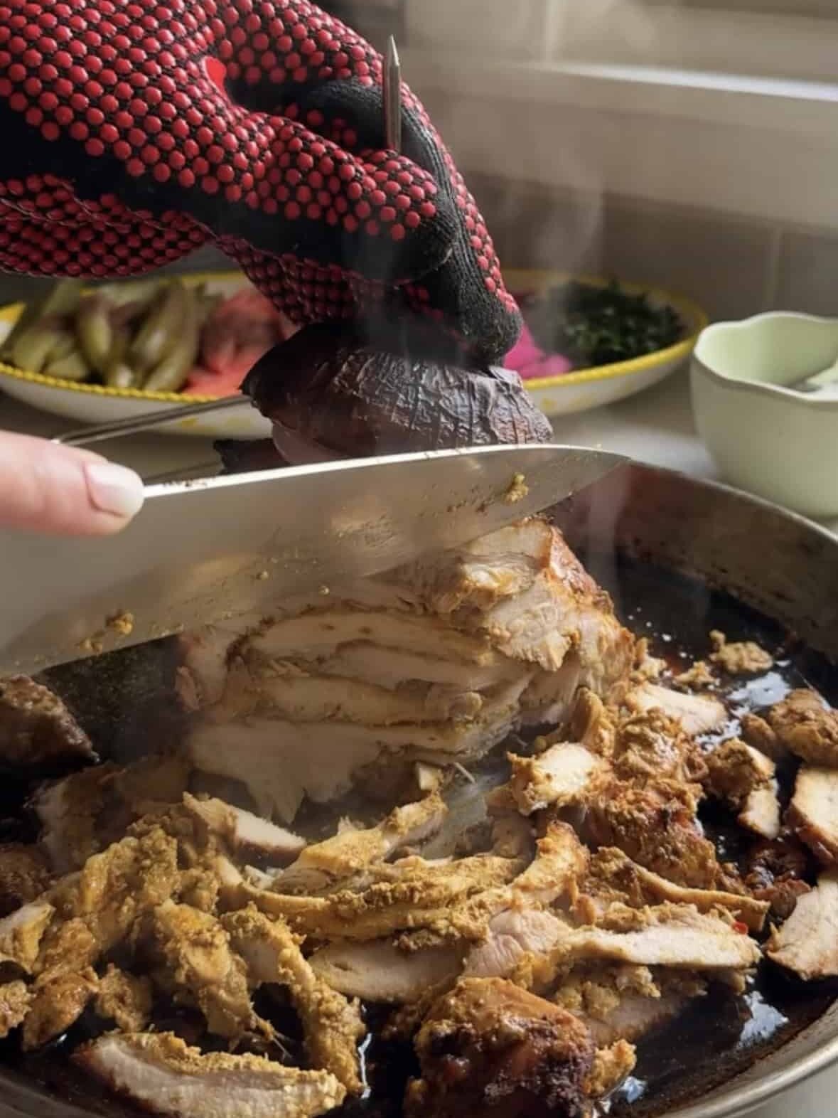 Slicing oven baked chicken shawarma off a vertical spit using a knife with oven mitt holding the spit steady and steam rising from the meat