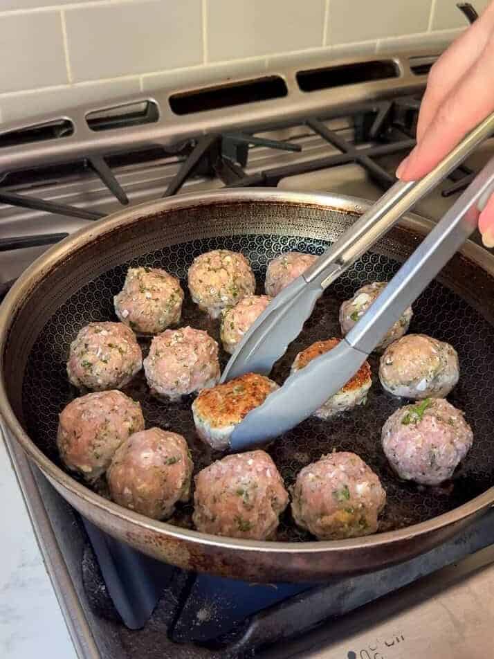 Searing Greek chicken meatballs in a skillet with tongs until golden brown
