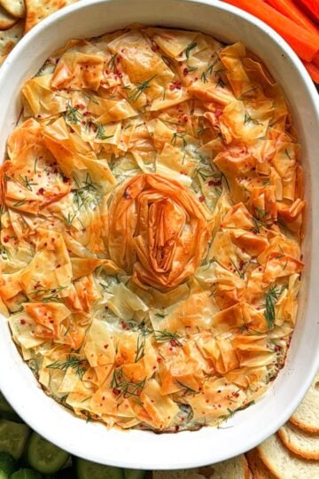 Baked spanakopita dip with a golden phyllo spiral, served with crackers, carrots, cucumbers, and toasted baguette