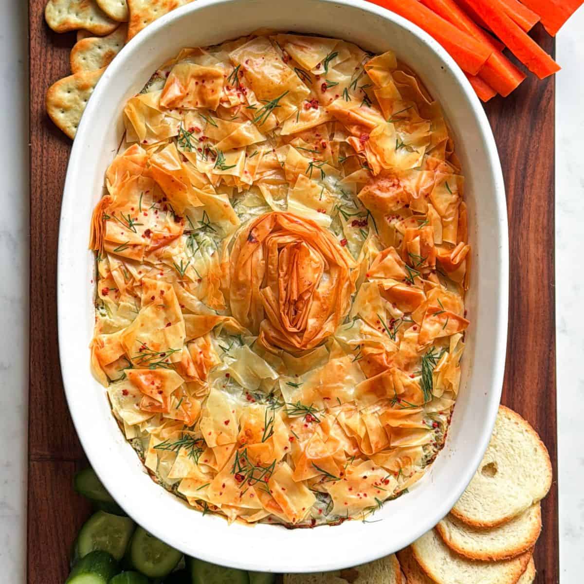 Baked spanakopita dip with a golden phyllo spiral, served with crackers, carrots, cucumbers, and toasted baguette