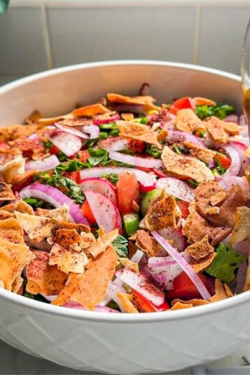 Pouring fattoush dressing over a bowl of fresh vegetables and toasted pita
