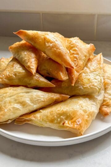 A plate piled high with golden baked cheese boreg triangles by a kitchen window