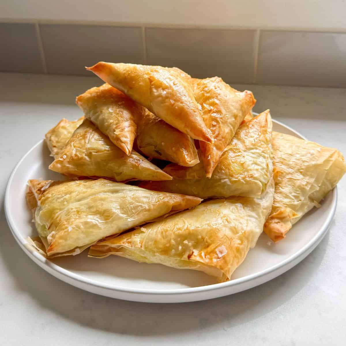 A plate piled high with golden baked cheese boreg triangles by a kitchen window