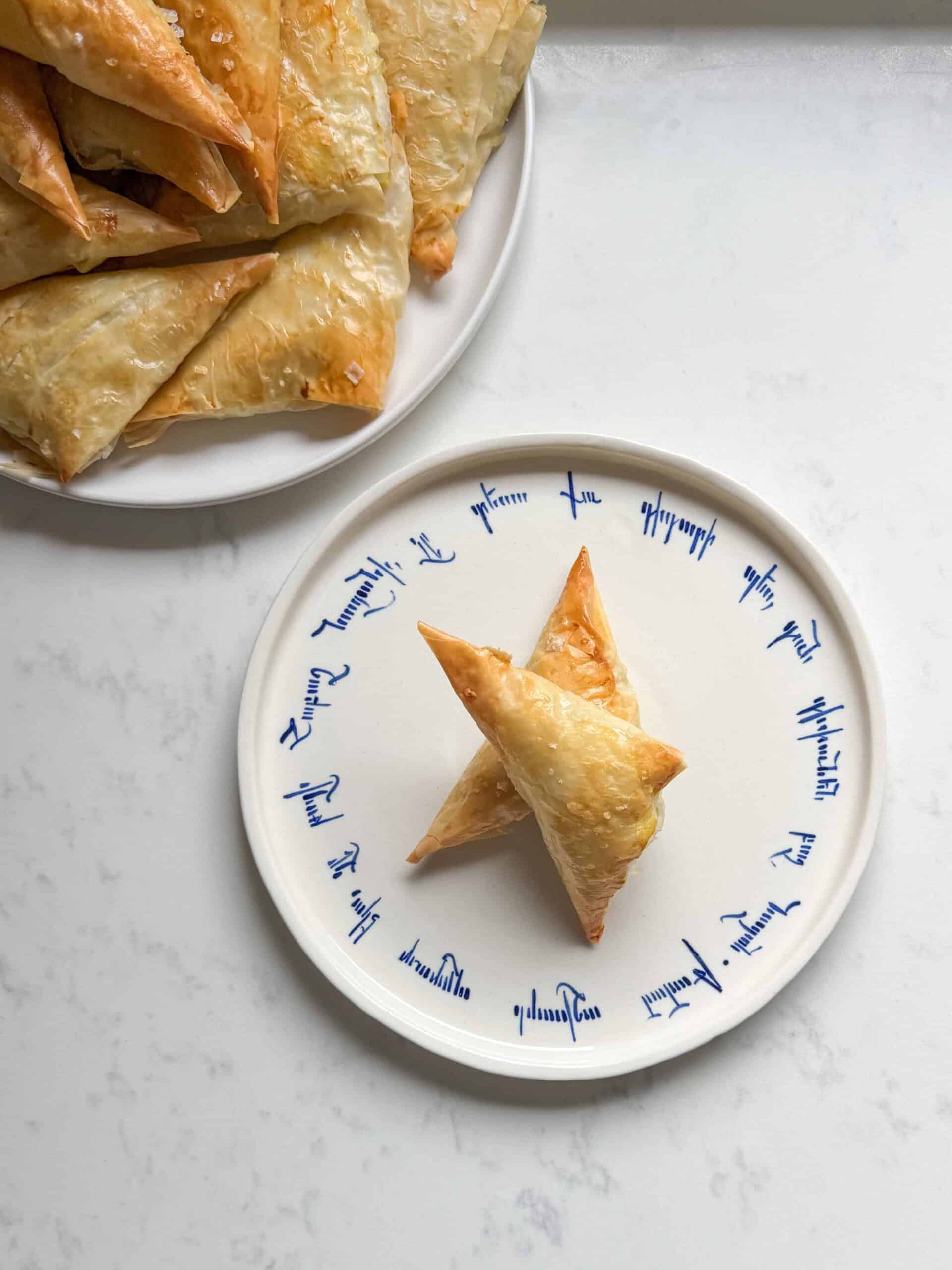 Baked cheese boreg triangles piled on a white plate next to a single triangle on an Armenian script plate