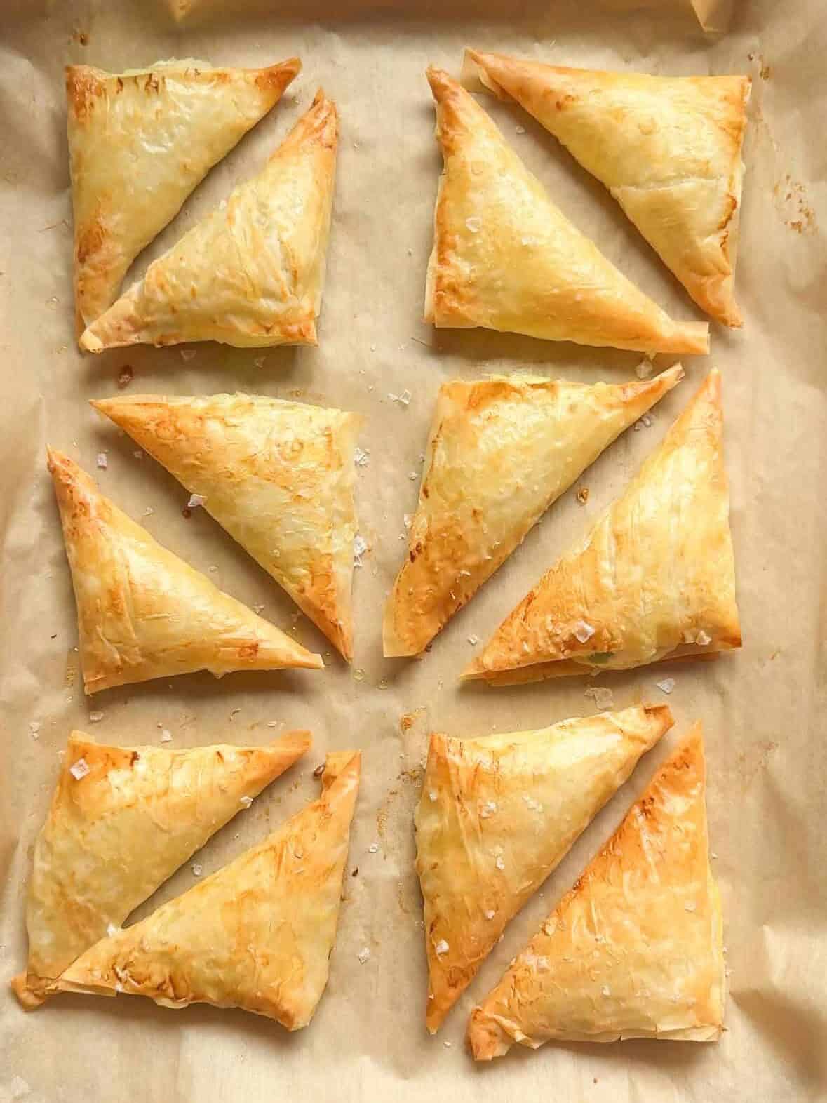 Overhead view of baked cheese boreg triangles arranged in rows on a parchment-lined baking sheet