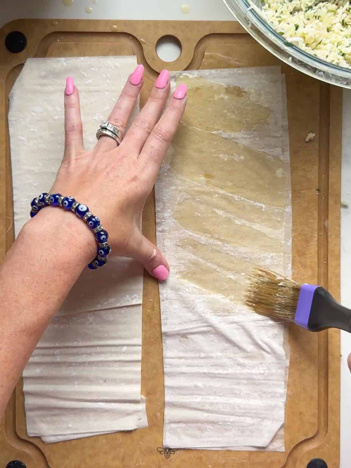 Hand holding phyllo dough flat while brushing a second sheet with melted butter on a cutting board