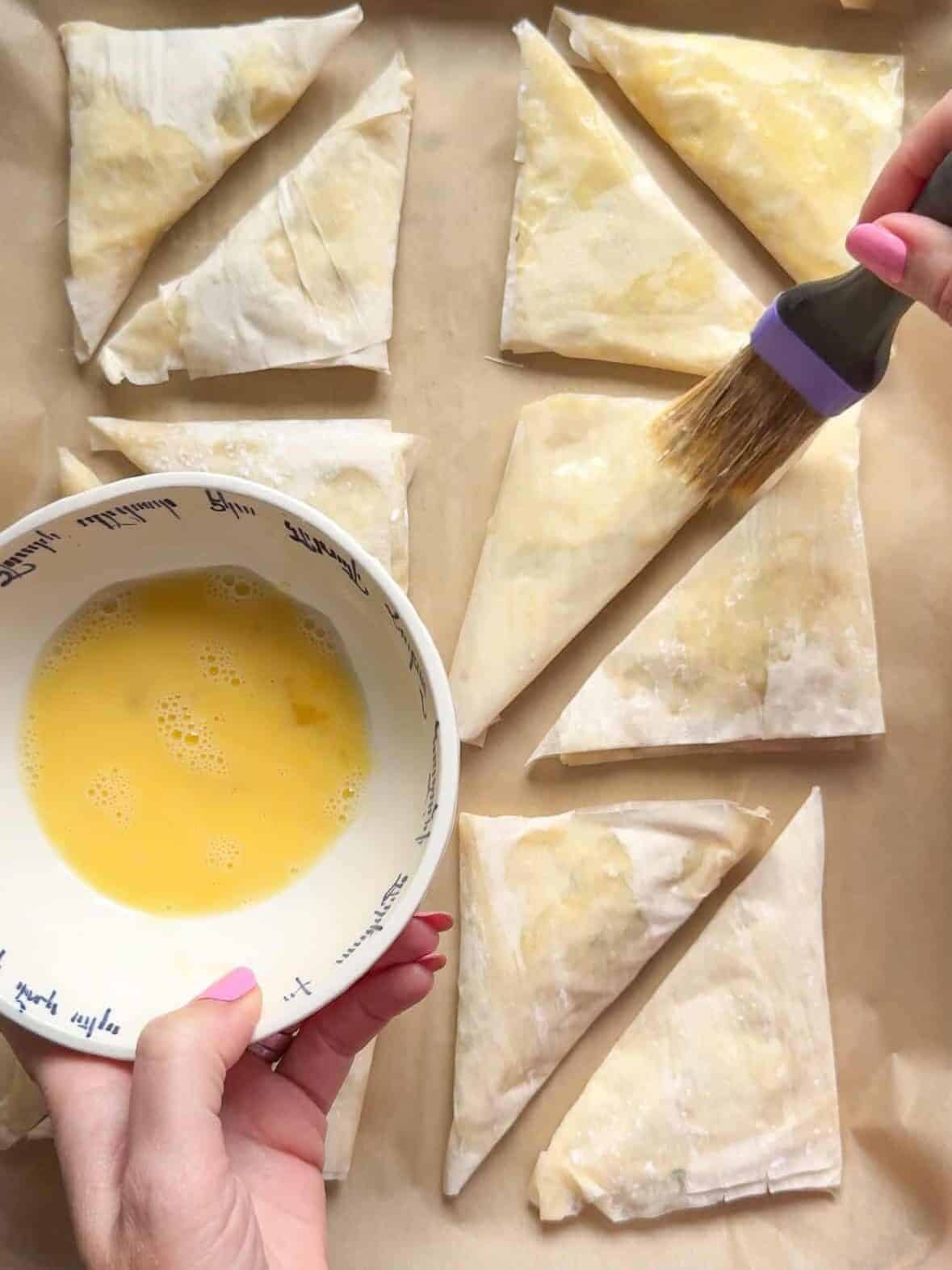Hand brushing egg wash onto unbaked cheese boreg triangles on a parchment-lined tray
