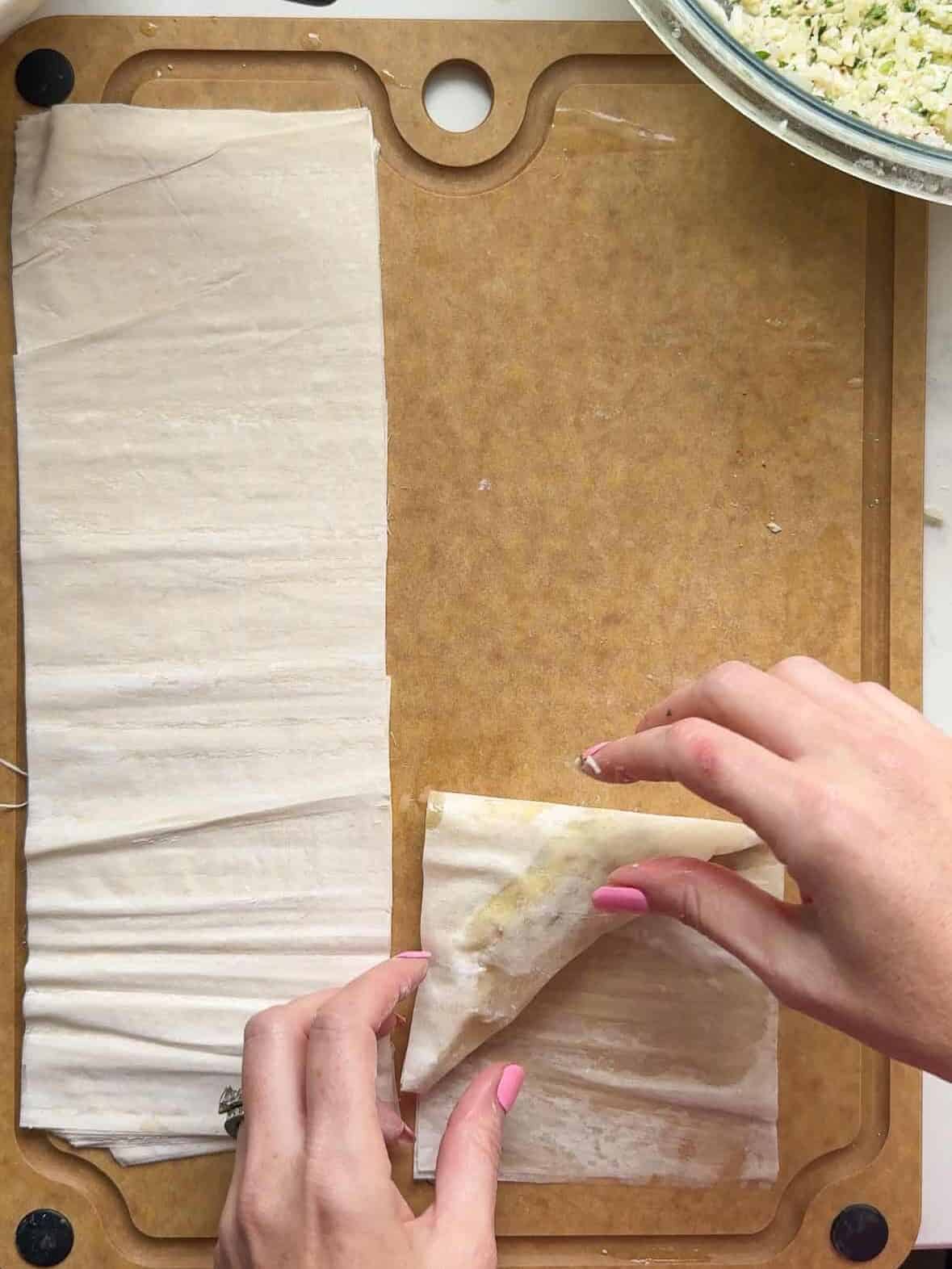 Hands folding a strip of buttered phyllo dough into a triangle for cheese boreg
