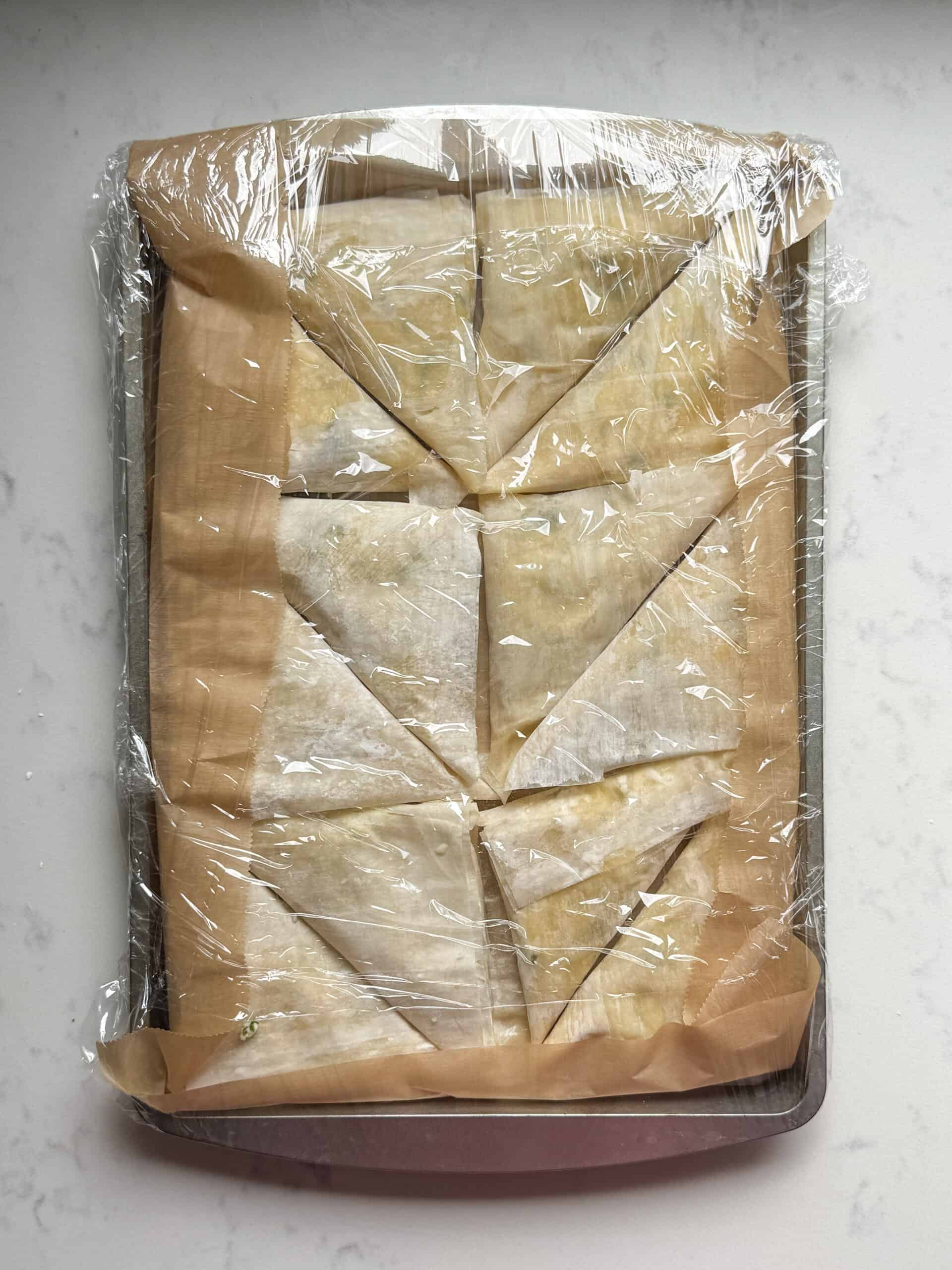 Unbaked cheese boreg triangles wrapped in plastic wrap on a tray before freezing