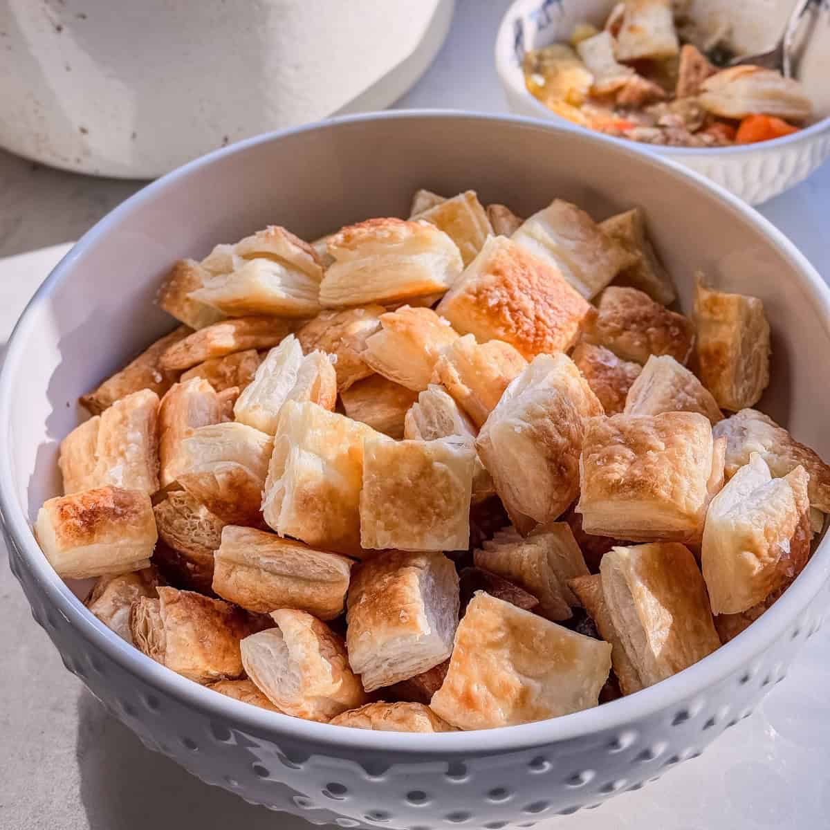 White bowl piled with puff pastry croutons, with a bowl of stew in the background