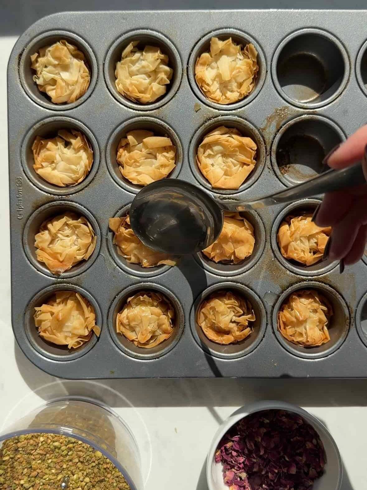 Spooning honey syrup over baked golden phyllo nests in a mini muffin tin, with bowls of crushed pistachios and dried rose petals.