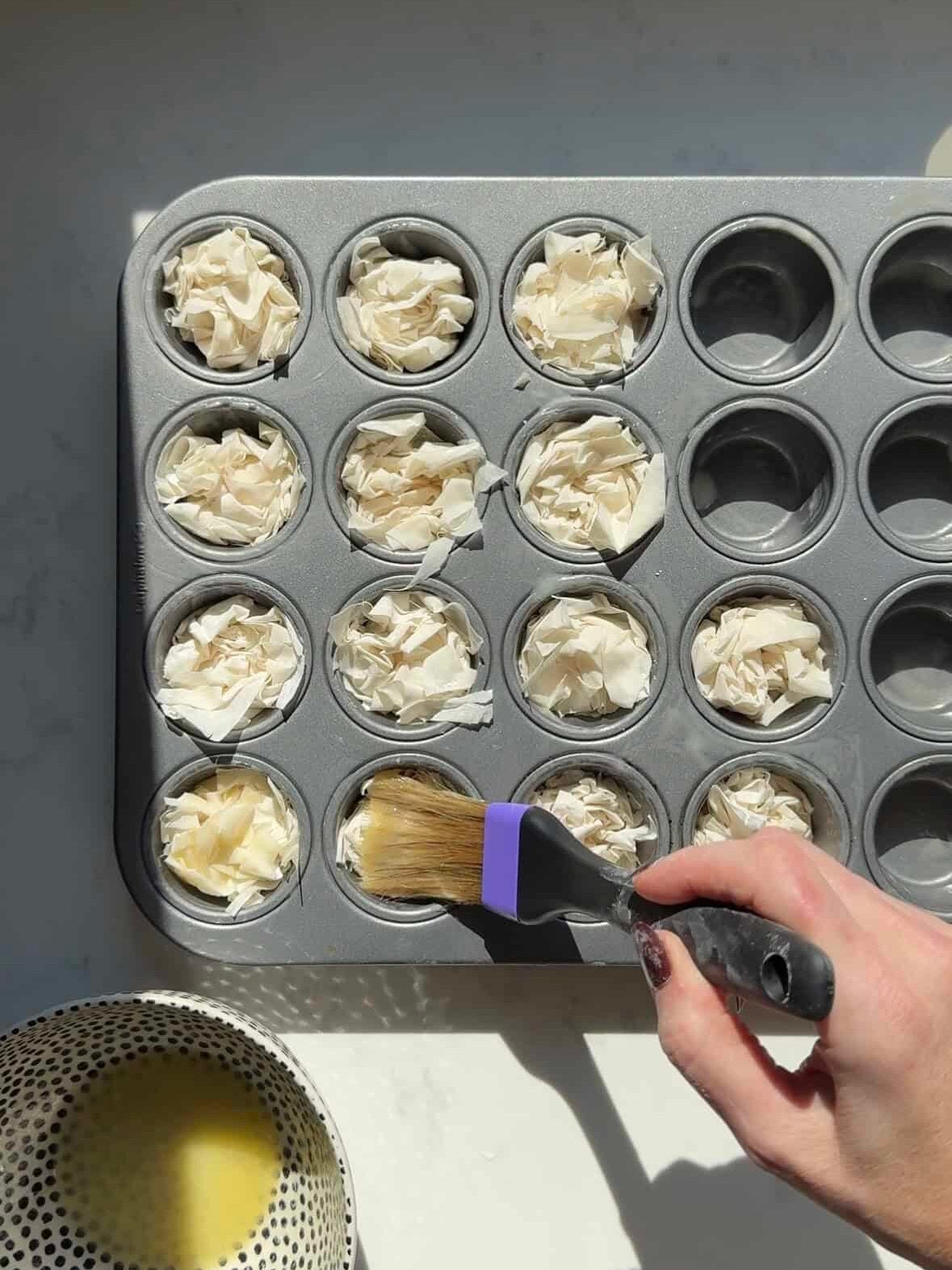 Crinkled phyllo cups in a mini muffin tin being brushed with melted butter.