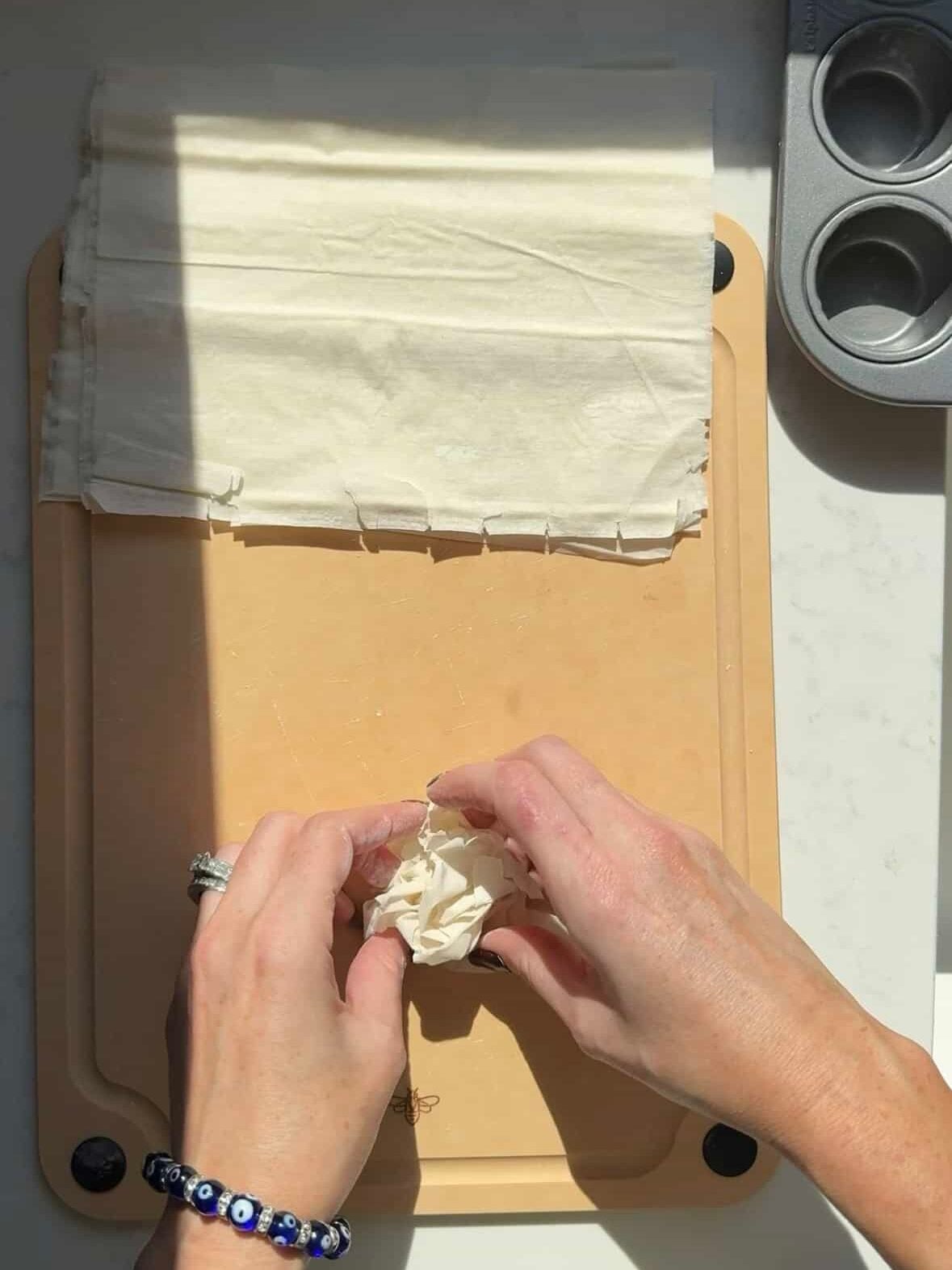 Hands crinkling a half sheet of phyllo dough on a cutting board next to a mini muffin tin.
