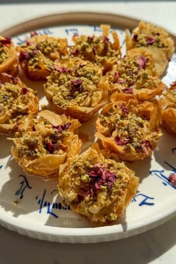 Close angled view of crispy golden phyllo baklava nests topped with crushed pistachios and rose petals.