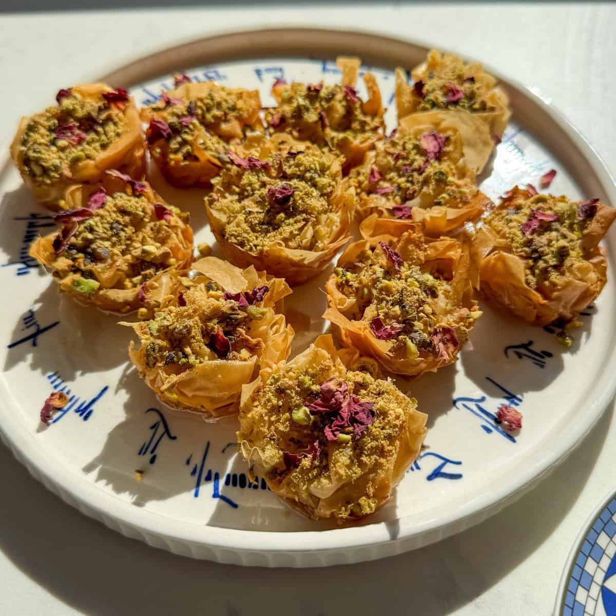 Close angled view of crispy golden phyllo baklava nests topped with crushed pistachios and rose petals.