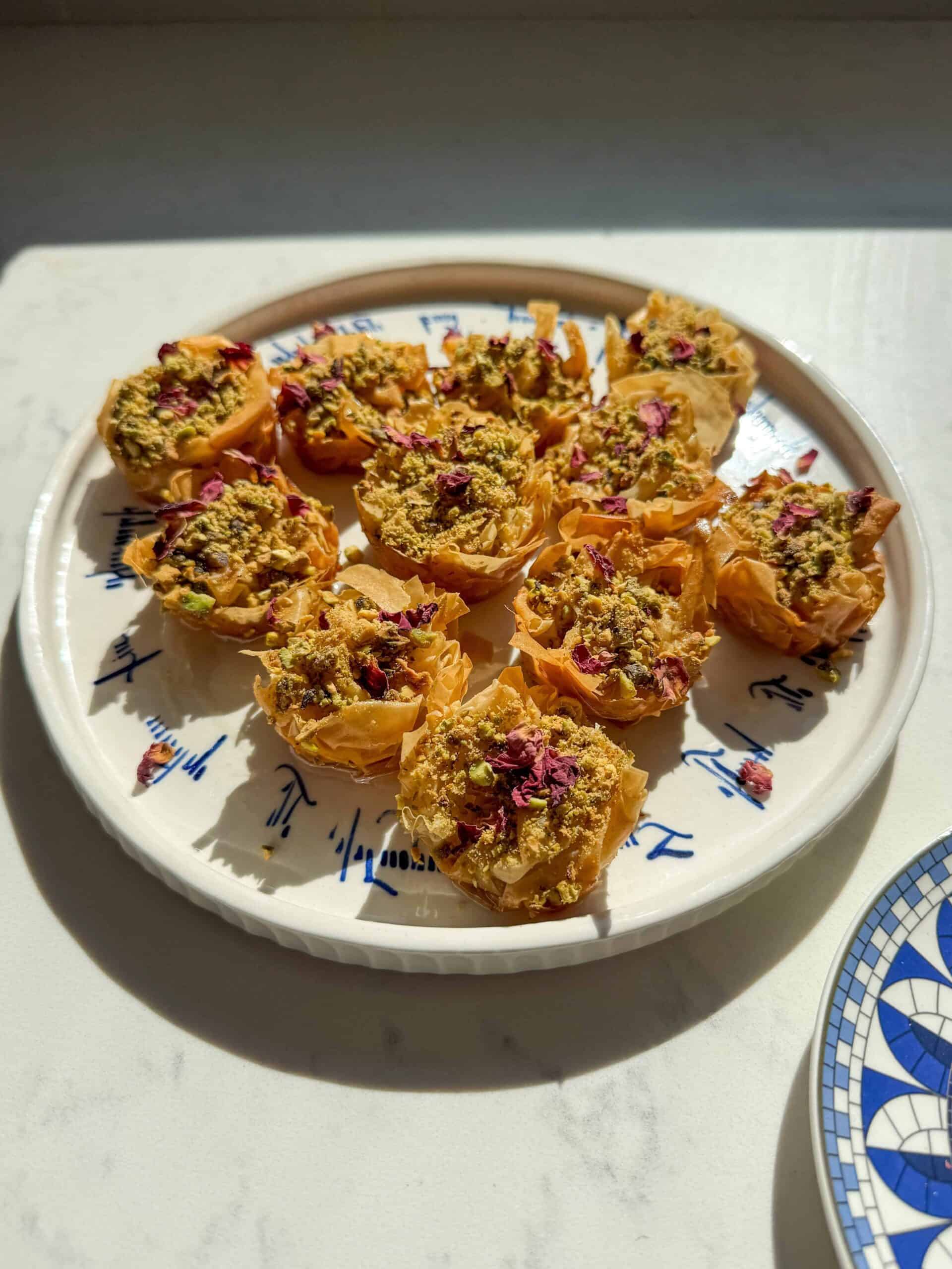 Close angled view of crispy golden phyllo baklava nests topped with crushed pistachios and rose petals.