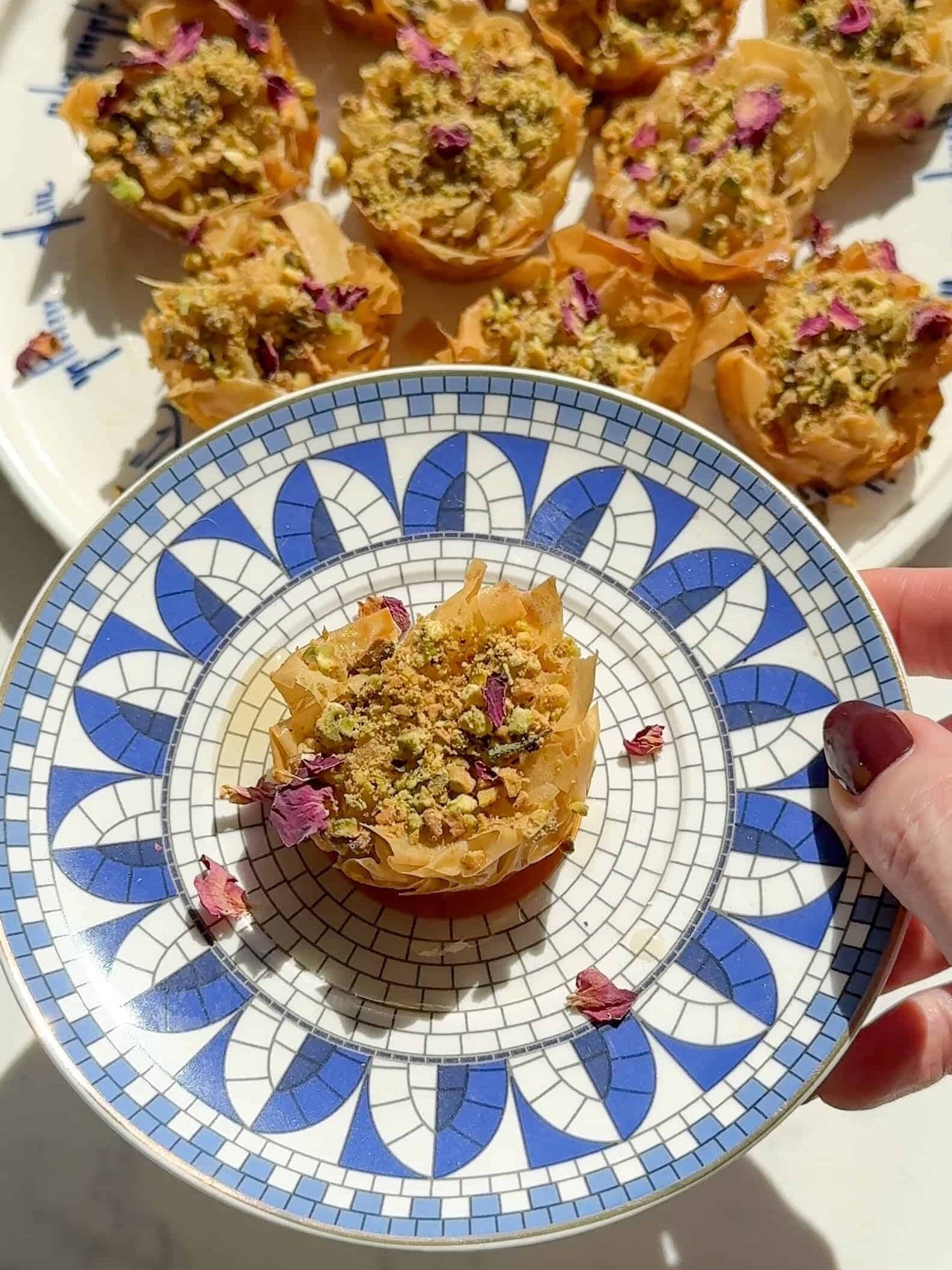 A single pistachio baklava nest with rose petals on a blue and white mosaic plate, with a platter of nests behind it.