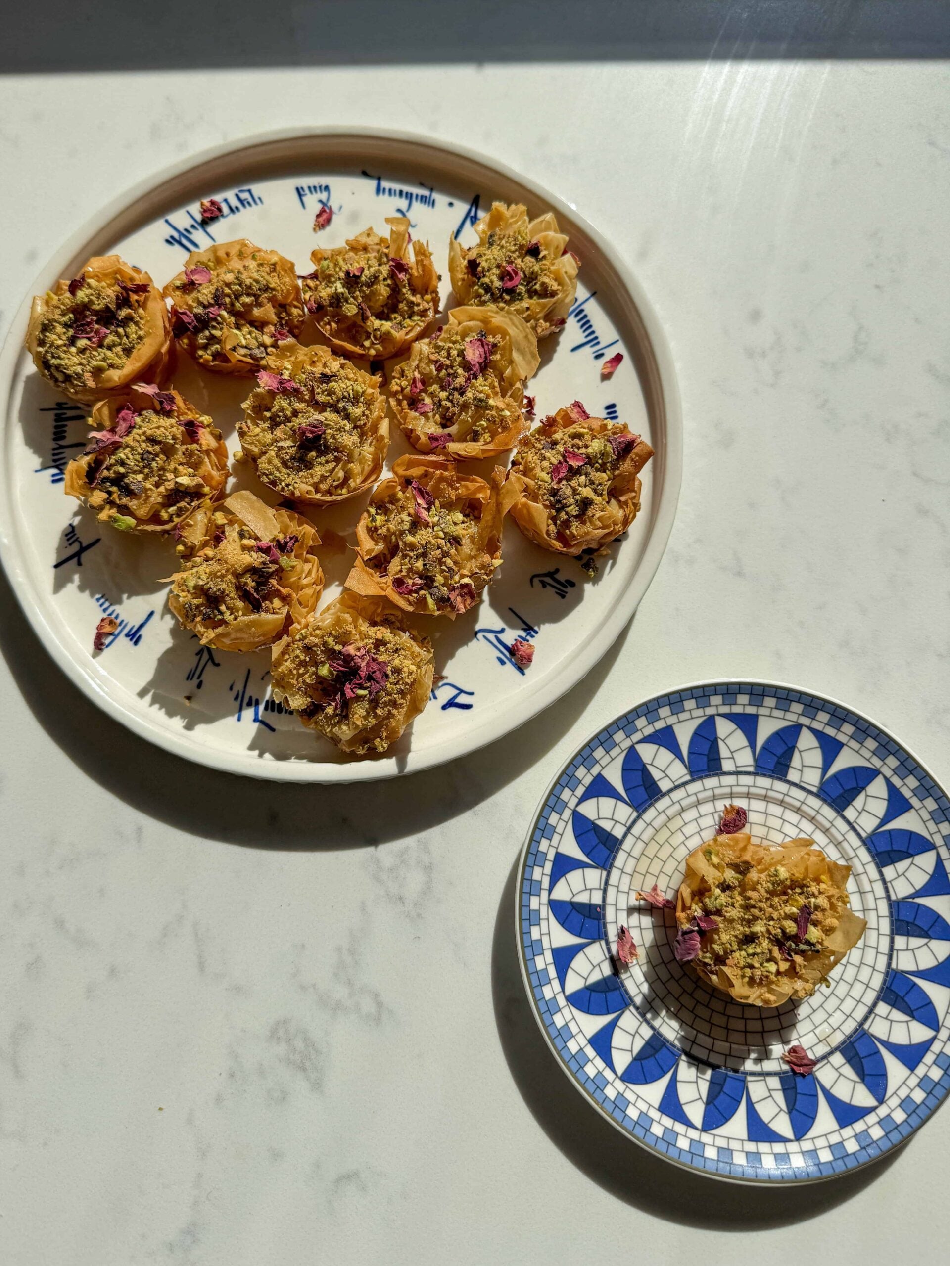 A platter of baklava nests beside a single nest on a blue and white mosaic plate.
A platter of baklava nests beside a single nest on a blue and white mosaic plate.