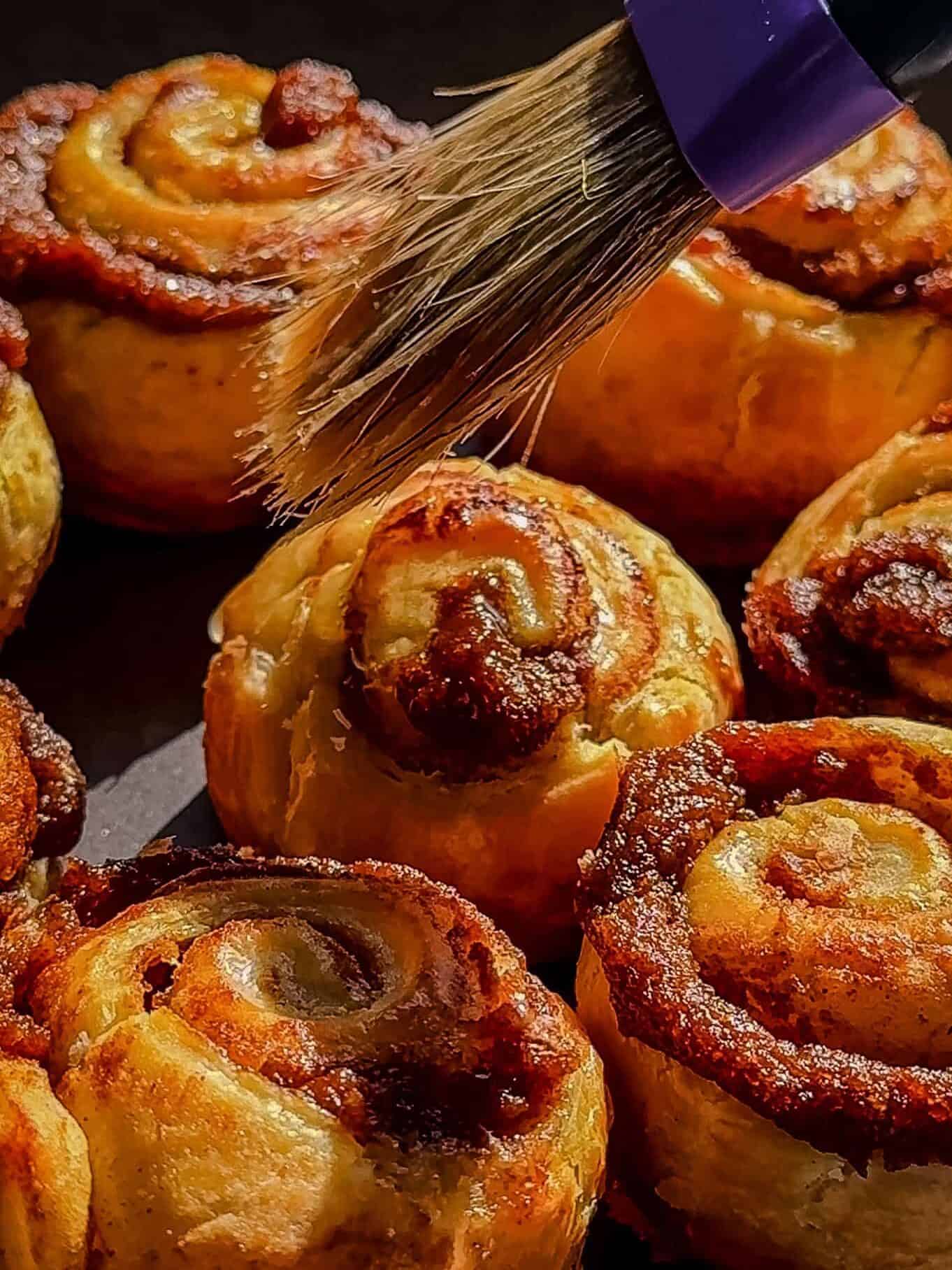 Brushing melted butter on baked pumpkin cinnamon rolls before coating them in cinnamon sugar
