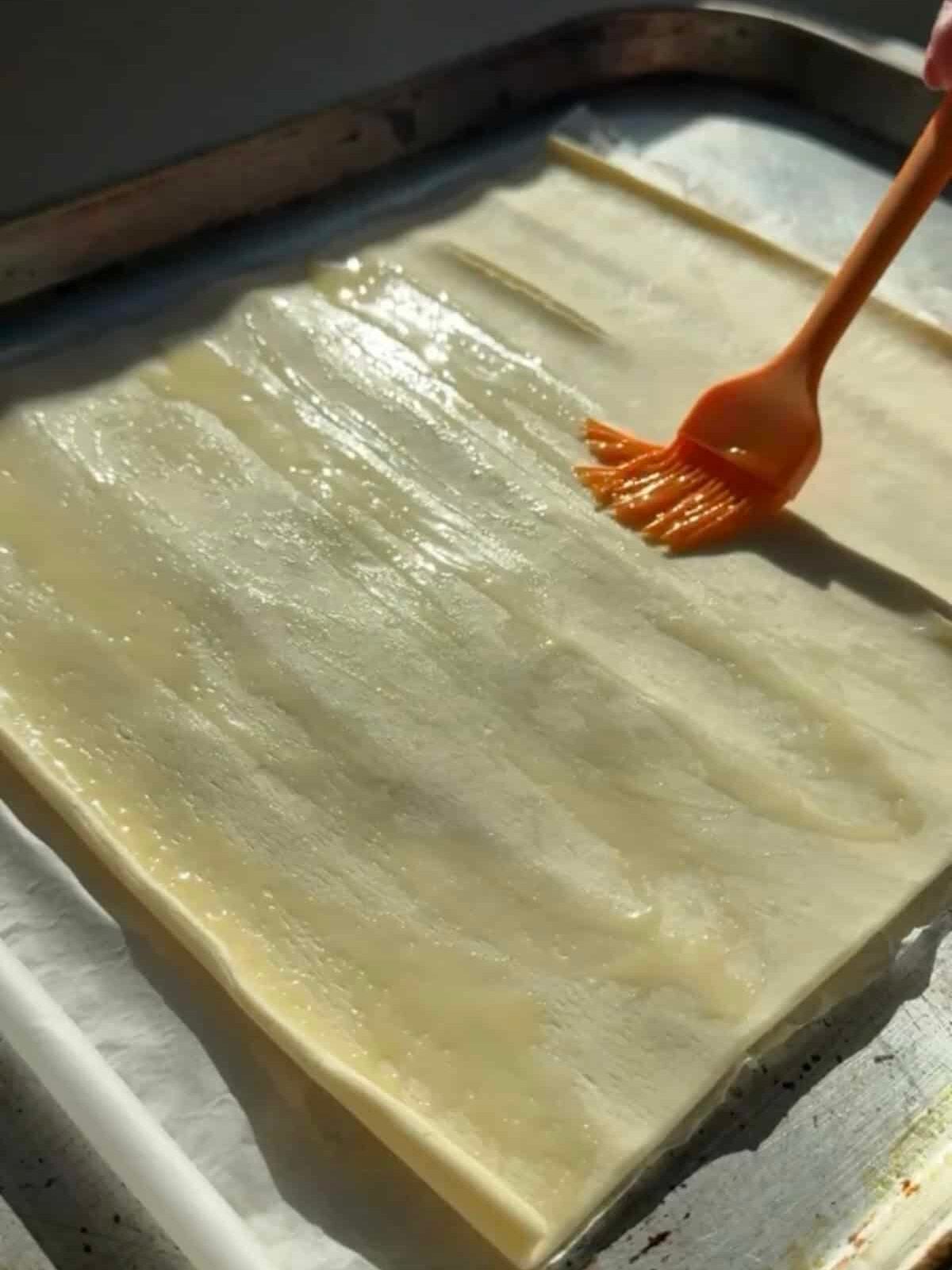 Pastry brush spreading melted butter over a sheet of puff pastry on parchment paper