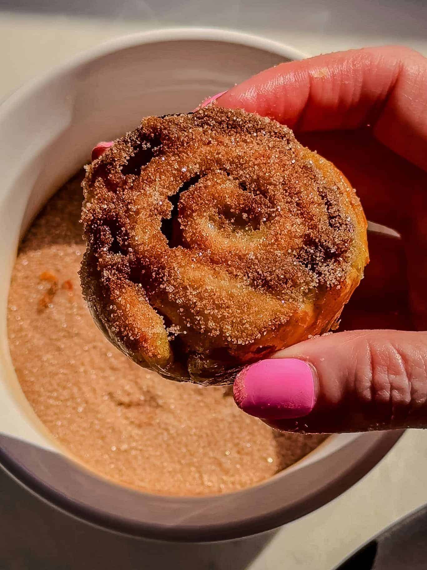 Hand holding a puff pastry cinnamon roll coated in cinnamon sugar over a bowl of cinnamon sugar