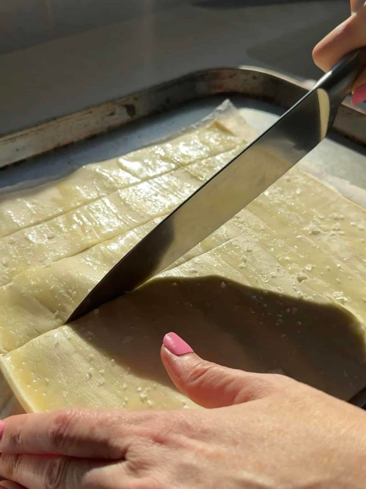 Knife cutting buttered puff pastry sprinkled with flaky sea salt into squares on a sheet pan