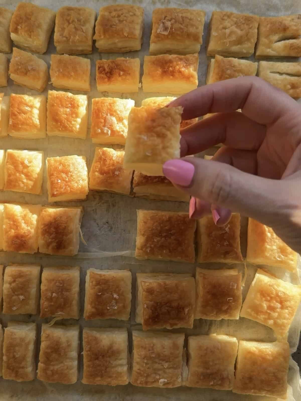 Hand holding a golden puff pastry crouton over a sheet pan of baked croutons